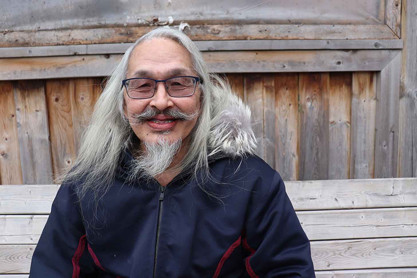 An older Inuit man with long gray hair and a long gray beard sits outside in a parka on a wooden bench