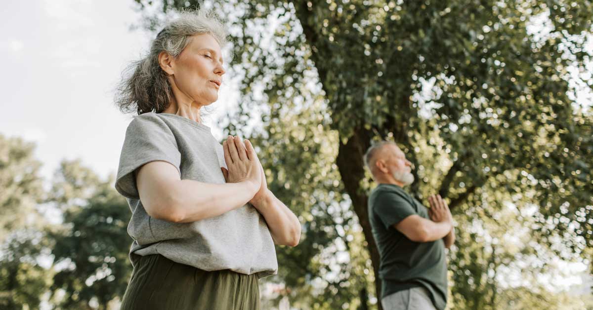 An elderly woman and man hold their hands in a prayer position at their chests as they do yoga in a park