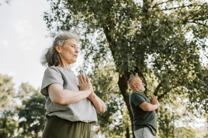 An elderly woman and man hold their hands in a prayer position at their chests as they do yoga in a park