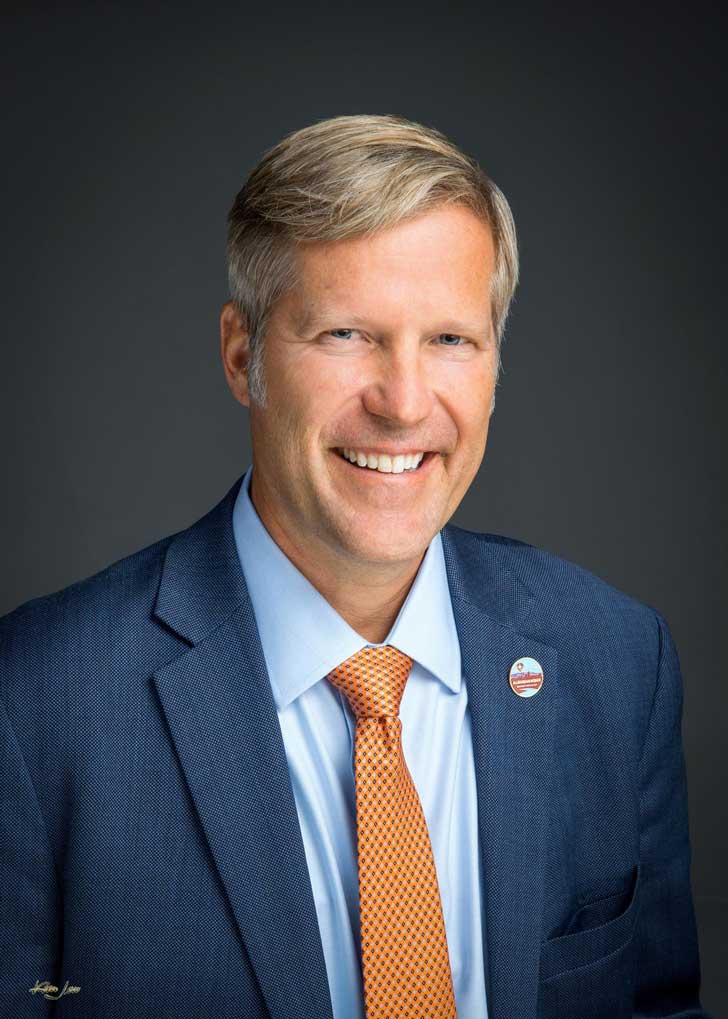 A headshot of Albuquerque Mayor Tim Keller, a white man with blond-gray hair, in a navy suit with an orange tie.