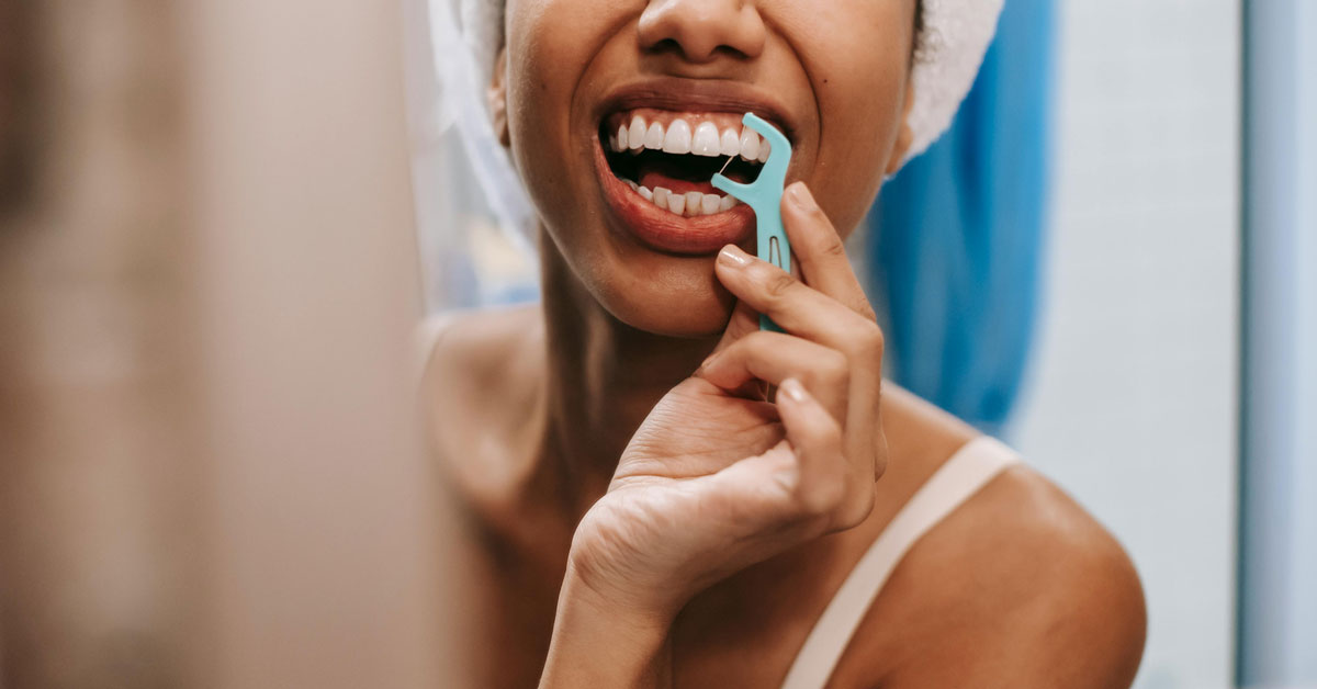 A close-up of a woman with melanated skin flossing her teeth with a floss pick in a bathroom mirror