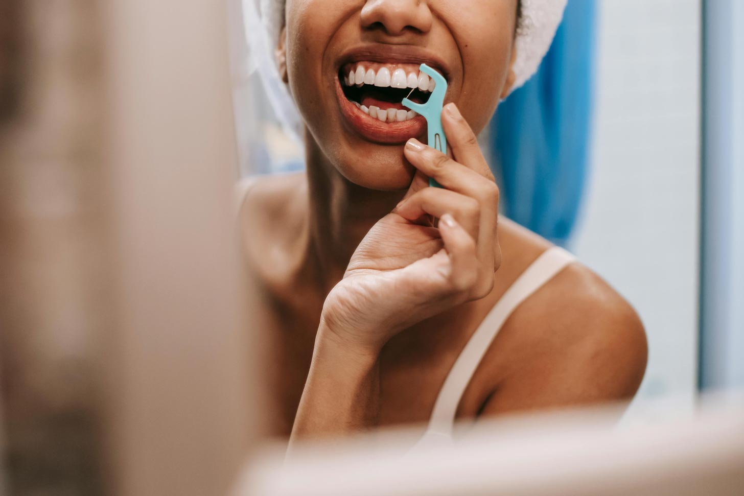 A close-up of a woman with melanated skin flossing her teeth with a floss pick in a bathroom mirror