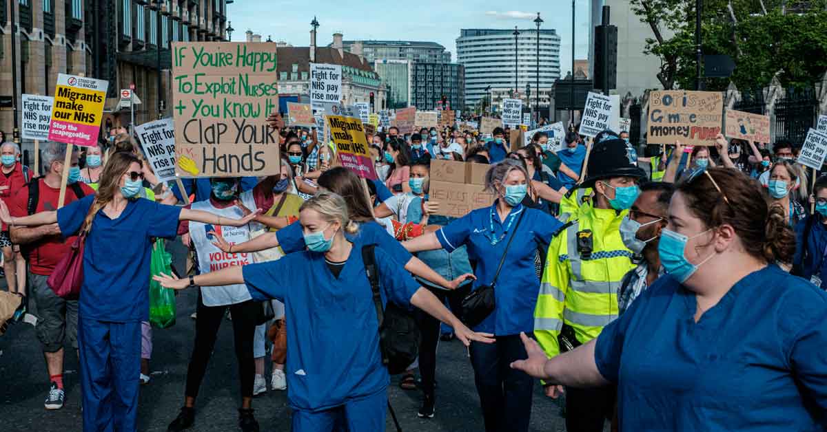 A nurse's union strikes in the streets, wearing blue scrubs and holding up protest signs