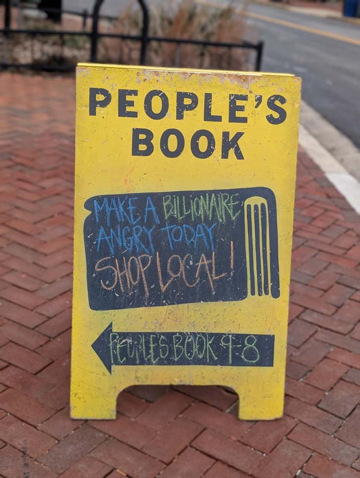 A yellow wooden sign outside of a store reads: "People's Book: Make a billionare angry today, shop local!" 