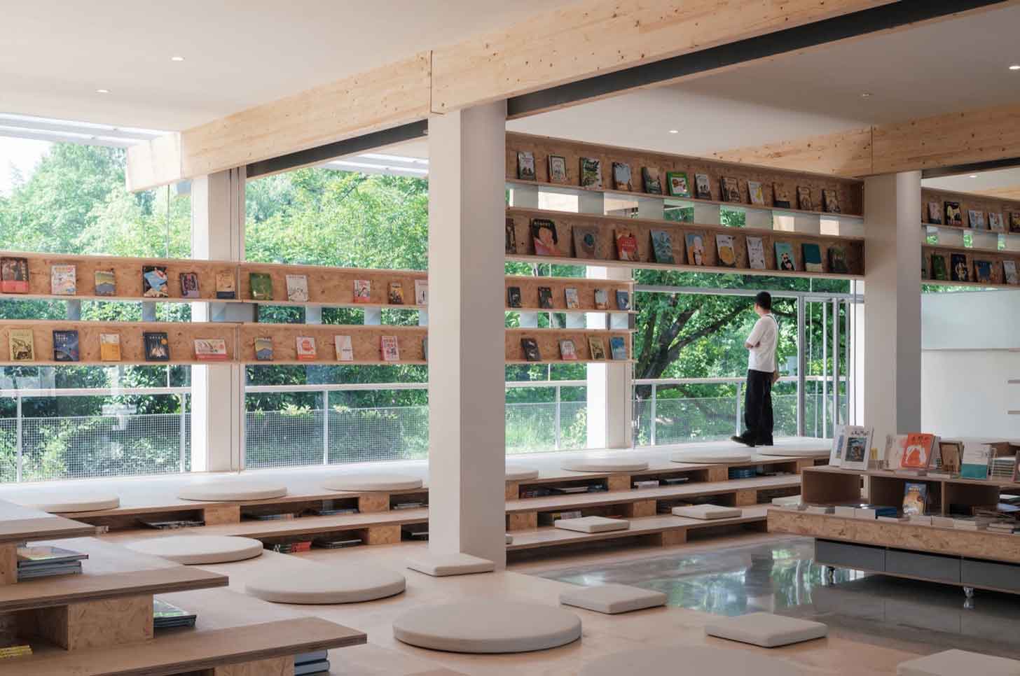 A bookstore customer looks out over a luscious green landscape through large windows, surrounded by bookshelves