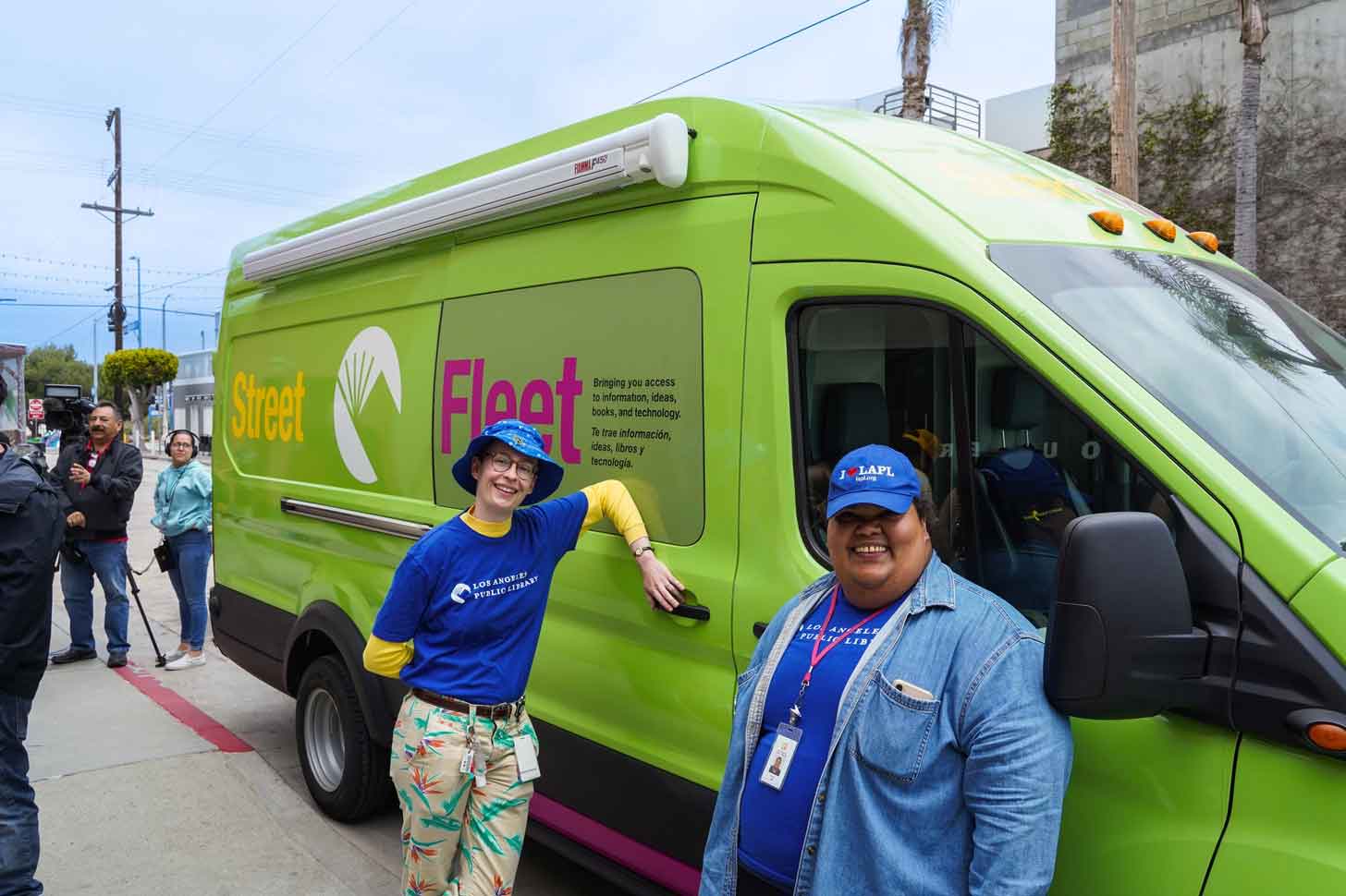 Two people wearing blue LA Public Library t-shirts stand outside of a green van that has a decal on the side, reading "Street Fleet." 
