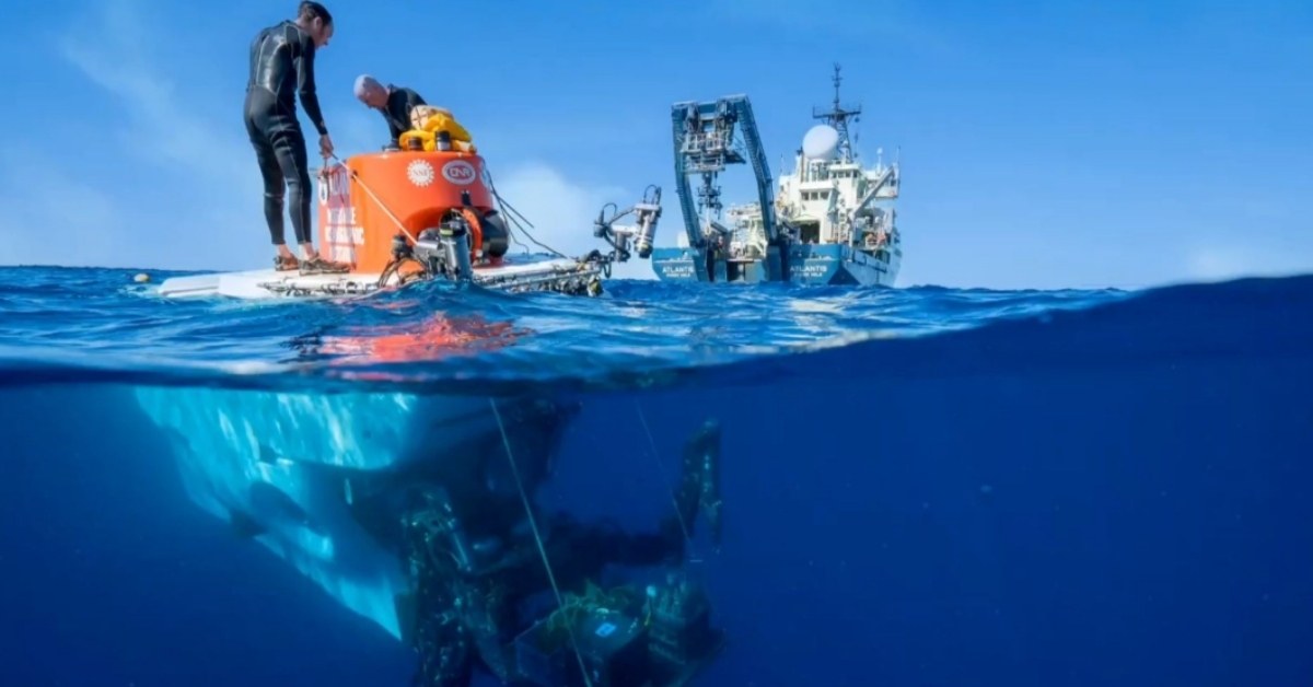 Two men stand atop a deep-sea submersible as it is launched just beneath the ocean's surface. A ship is in the background, as well as a blue sky