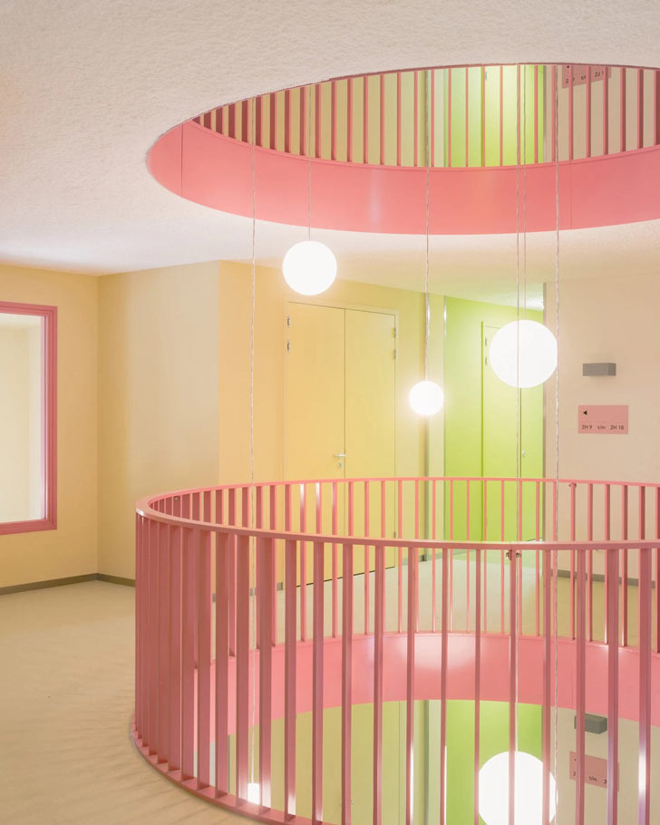 A hallway opens up to a pink circular staircase, with yellow and green accents, and spherical lighting