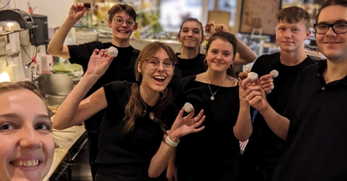 A group of six teenagers smile and take a selfie together inside a restaurant as they hold up small balls of dough.
