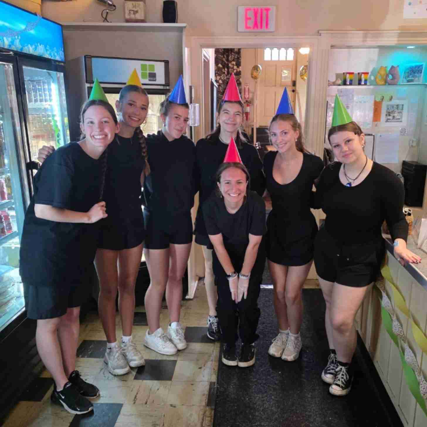 Seven teenage girls smile as they take a group photo together inside a restaurant, wearing party hats