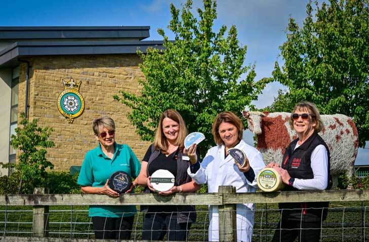 Four women stand in front of a tan brick building a cow statue as they hold up pieces of wrapped cheese and smile