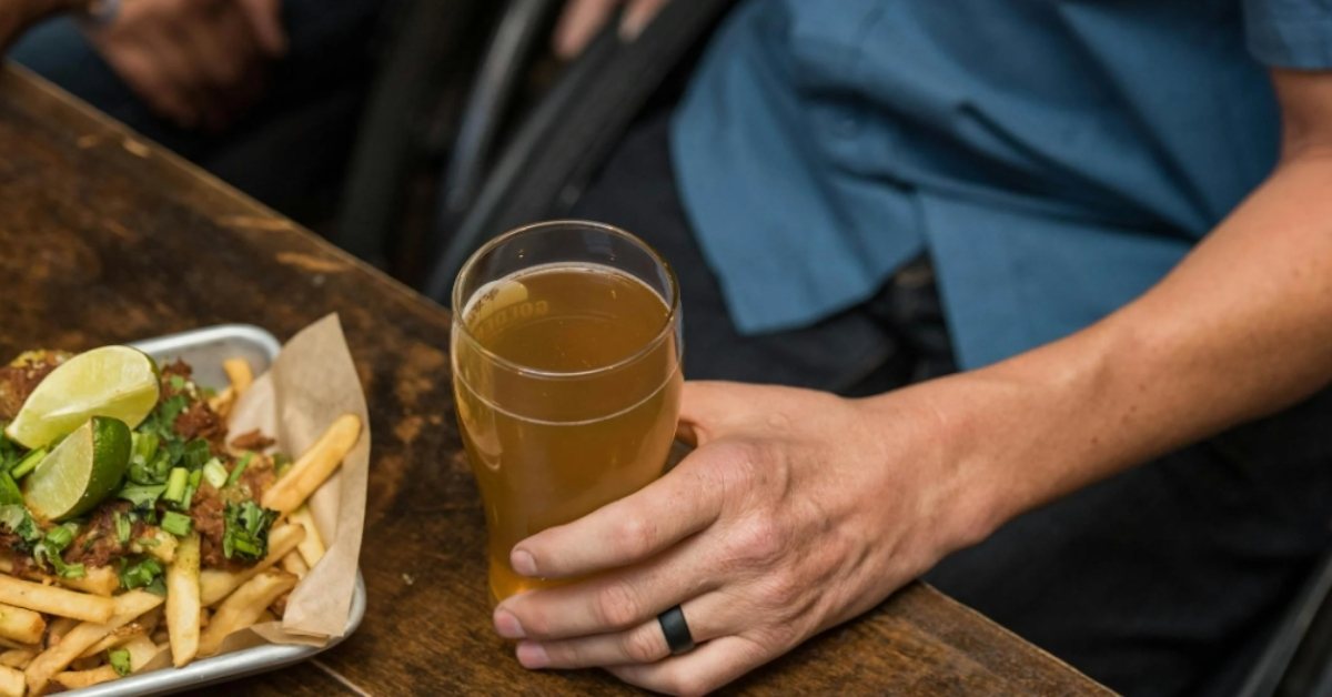 A wheelchair user drinks a beer at a table next to a box full of fries. 