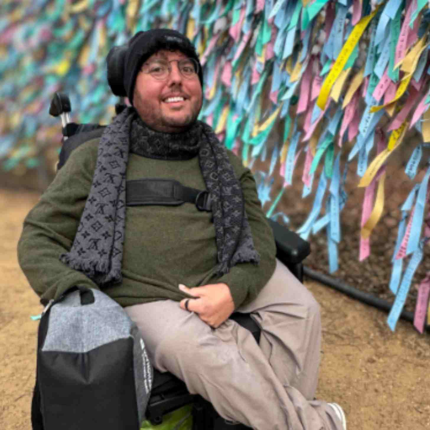 A man in a wheelchair sits in front of a wall filled with colorful pieces of paper where people have written wishes and prayers