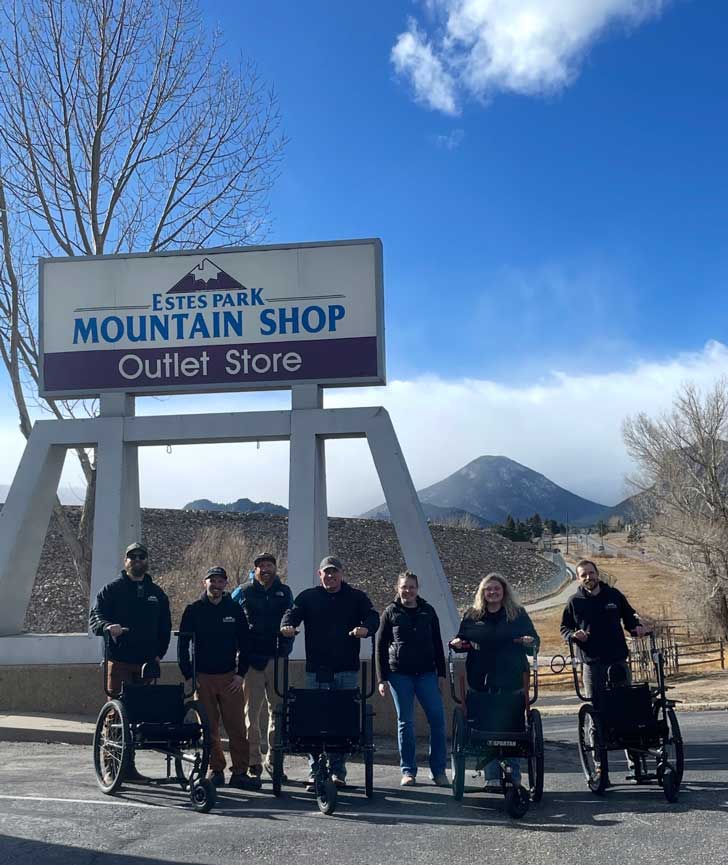 Seven people stand outside of the Estes Park Mountain Shop in Rocky Mountain National Park, holding four all-terrain wheelchairs