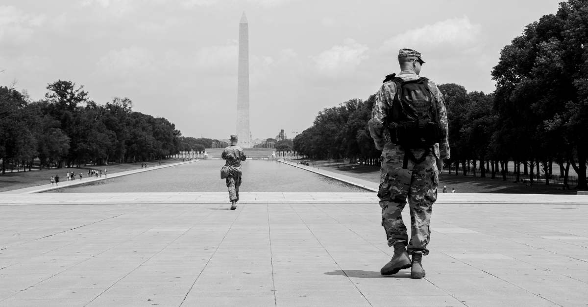 A man wearing an army uniform with a backpack on his back walks at a park