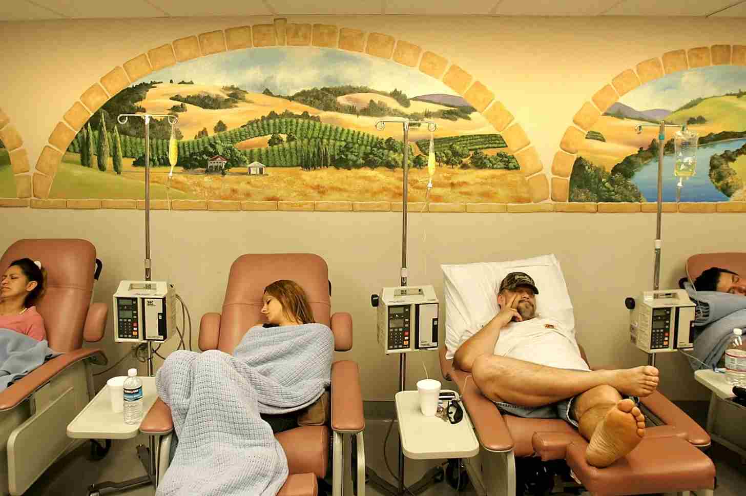 A row of people in recliners sit next to IV bags of antiobiotics in front of a mural depicting the Central Valley of California
