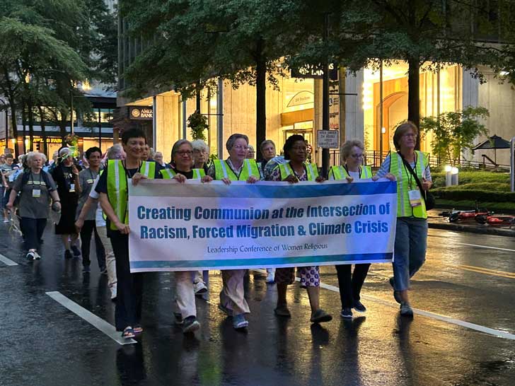 Six women wearing bright yellow traffic vests march down a downtown Atlanta street with a banner that reads "Creating Communion at the Intersection of Racism, Forced Migration & Climate Crisis"