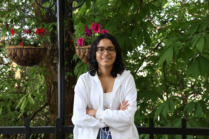 A 16-year-old girl with shoulder-length black hair and black glasses smiles in front of lush green plants and pink flowers
