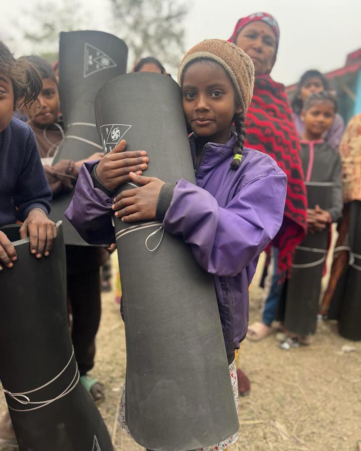 A small Nepalese girl in a purple coat and brown hat hugs a rolled-up black rubber mat, smiling gently