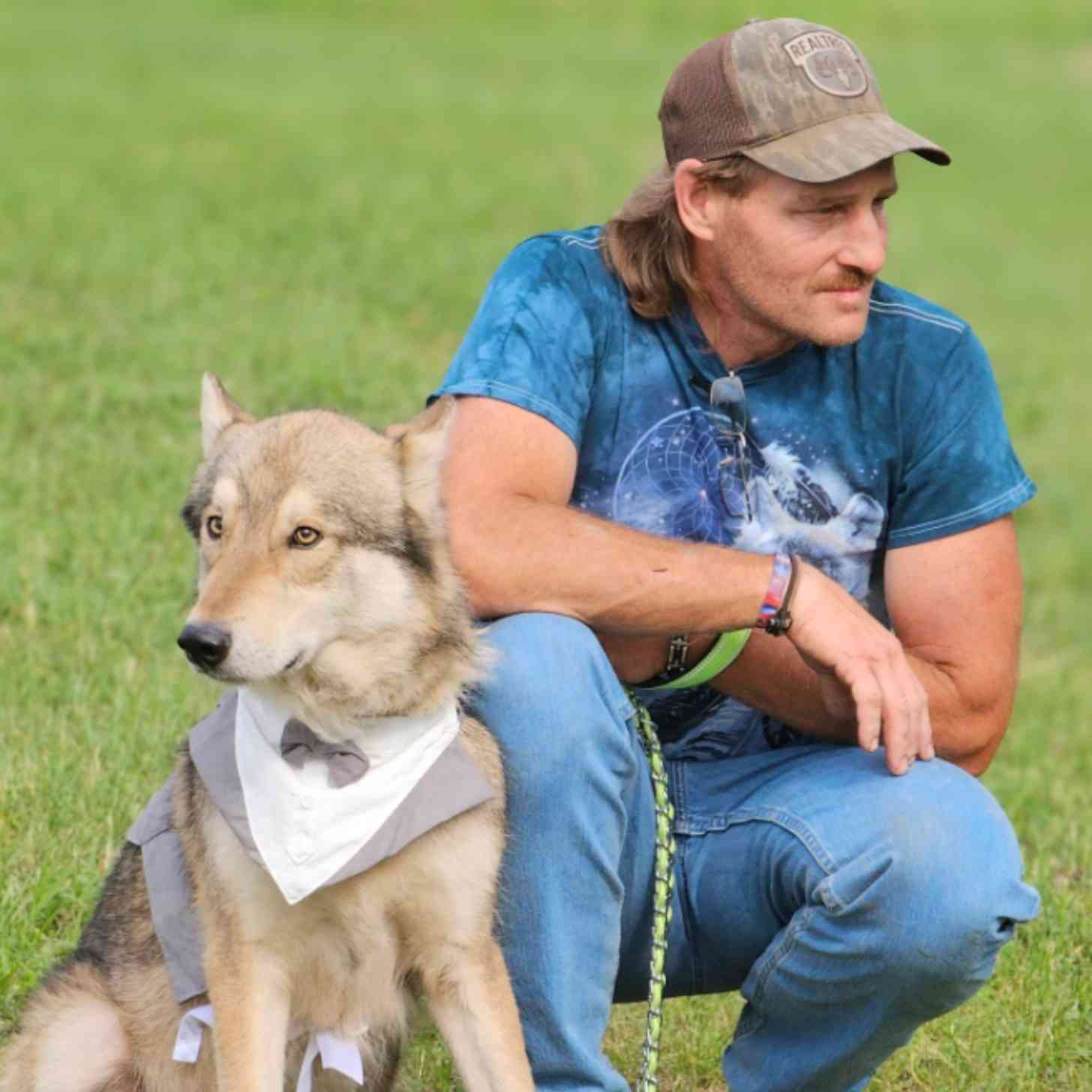 A man with a wolf shirt on kneels in a field beside a white wolfdog