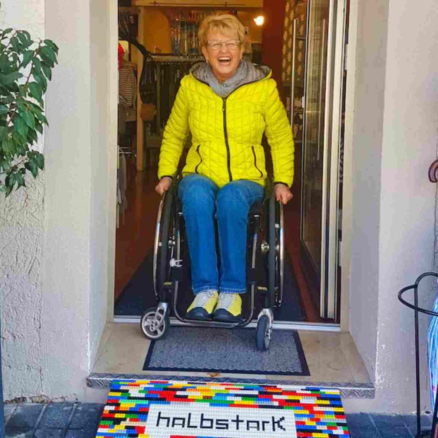 An elderly woman smiles as she sits at the top of  a staircase, with a lego wheelchair ramp below her