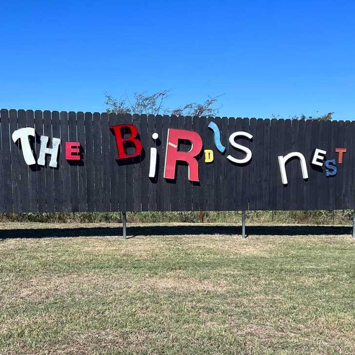 On a fence, metal letters of different colors and sizes spell out "The Bird's Nest"