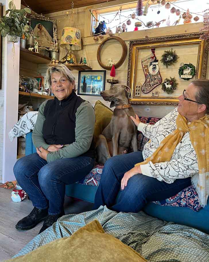 Two older women sit inside of a small home, which is covered in eclectic decor. Between them sits a grey dog