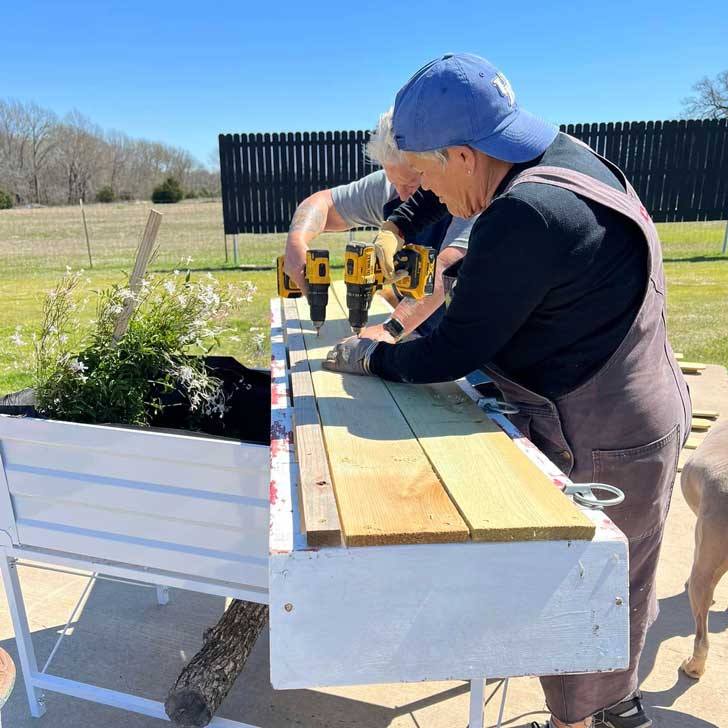 Two older women take a drill to a piece of plywood