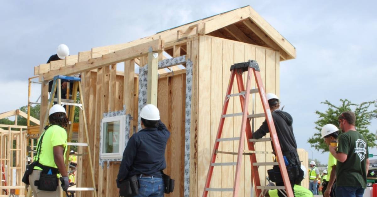 Teens in construction hats and vests surround a tiny home mid-build.
