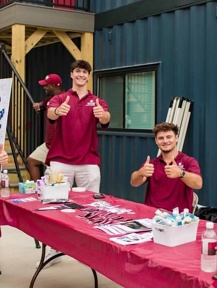 Two male residents stand outside of a shipping container dorm, giving thumbs up in red polos