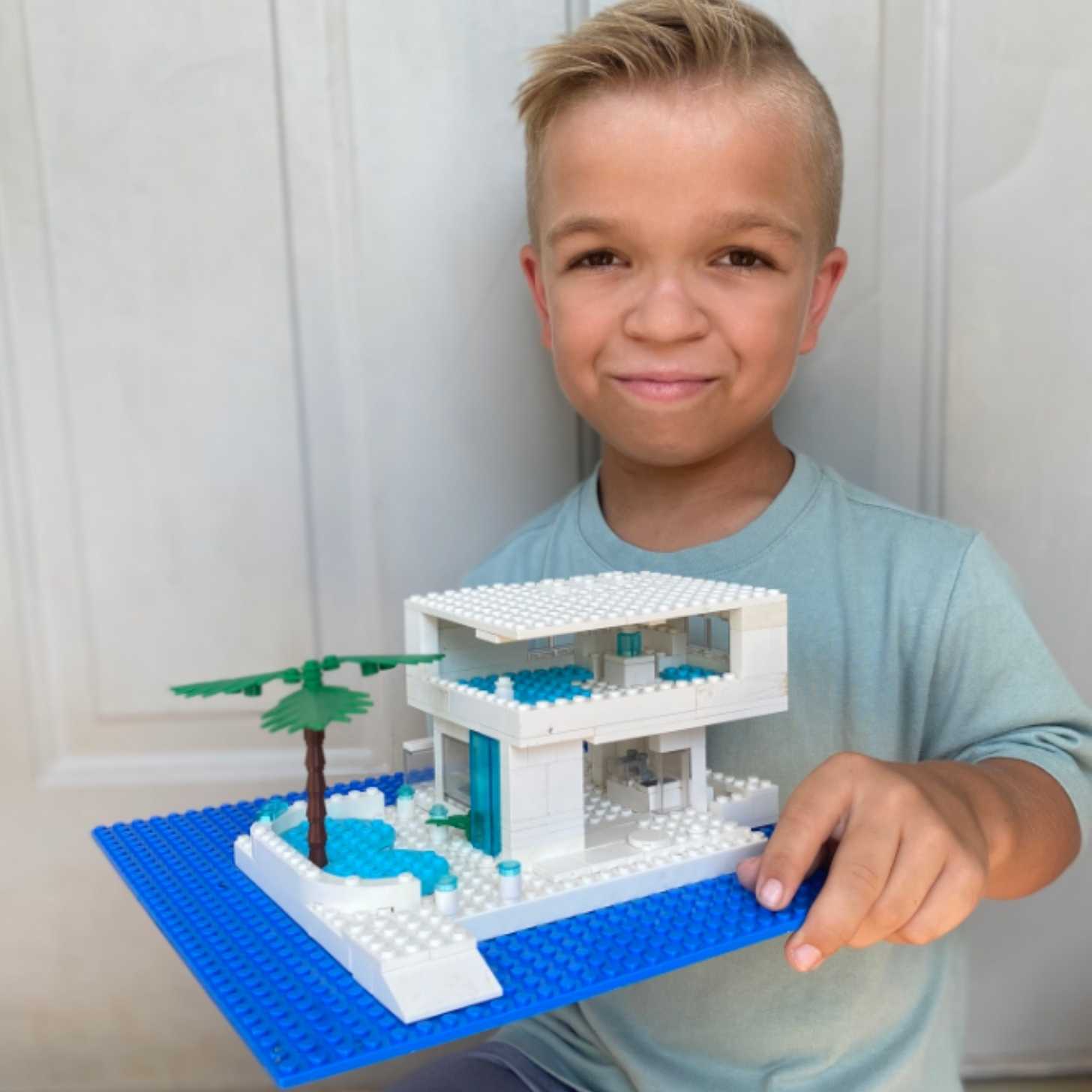 A young blonde boy smiles slightly as he holds up a LEGO house called Island Oasis Beach House.