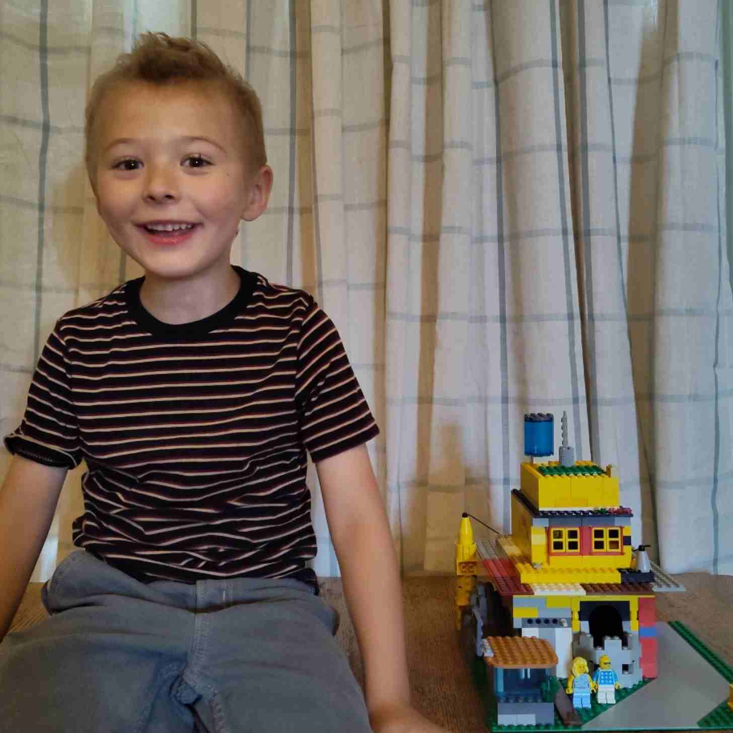 A little boy with stuck up hair smiles broadly as he sits on a bed beside his LEGO creation, titled Ssembler's House