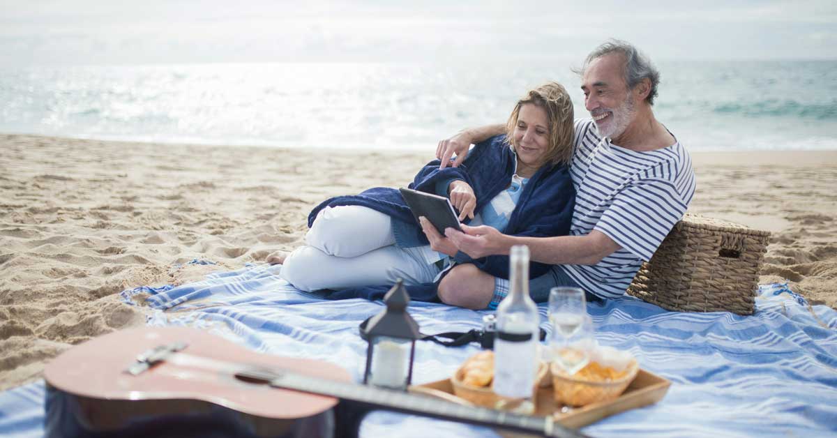 An elderly man and woman sit on a picnic blanket on the beach, next to a picnic basket and guitar
