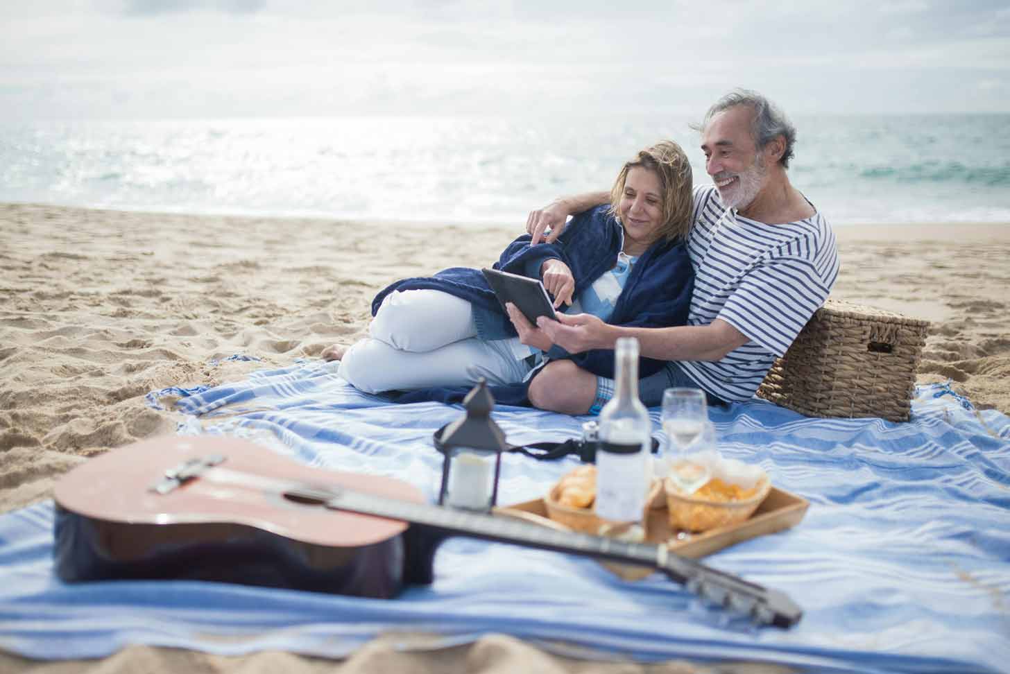 An elderly man and woman sit on a picnic blanket on the beach, next to a picnic basket and guitar