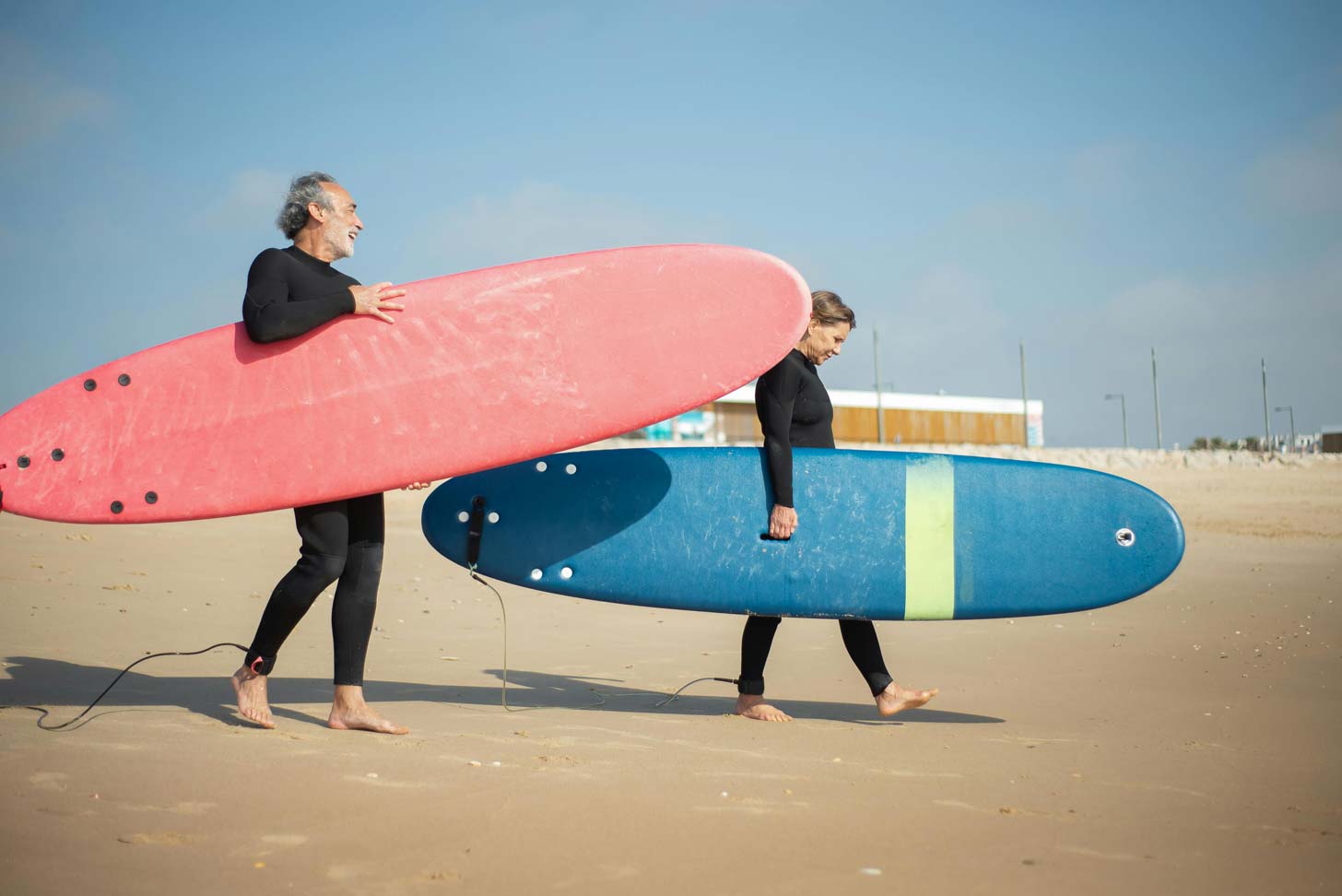An elderly couple wearing black wetsuits take pink and blue surfboards out to the water, walking on the beach