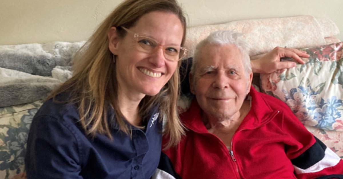 A woman with blond hair and clear glasses sits on a floral couch next to an elderly man in a red shirt