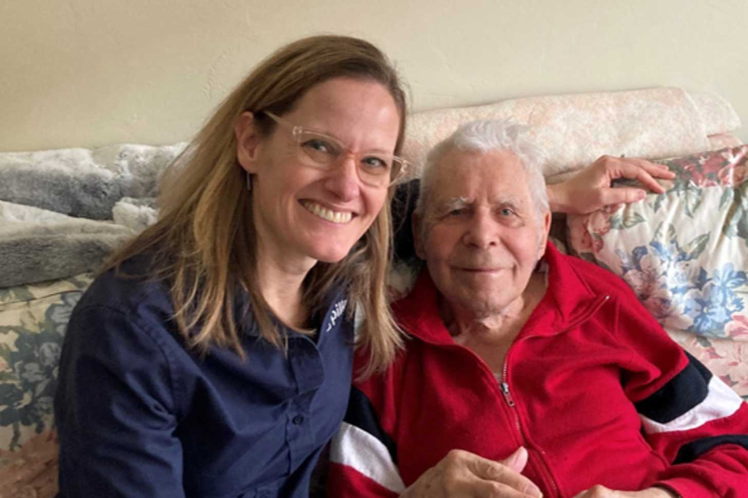 A woman with blond hair and clear glasses sits on a floral couch next to an elderly man in a red shirt