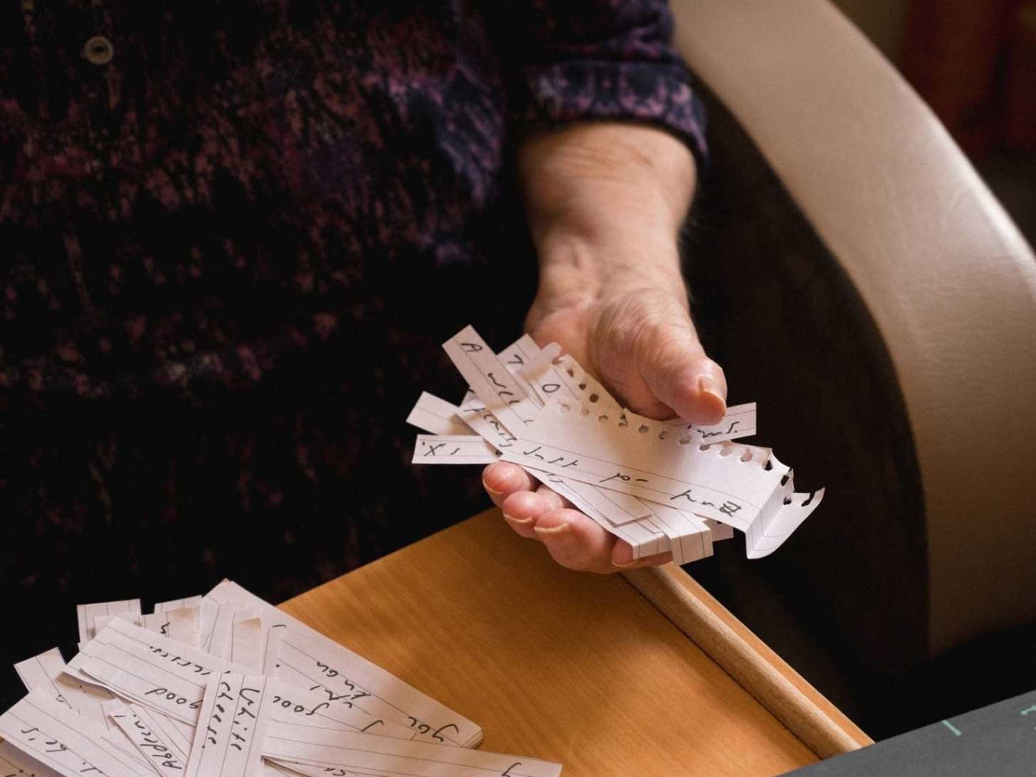 A close-up of an elderly woman's hand, holding scraps of paper with illegible writing scrawled on them