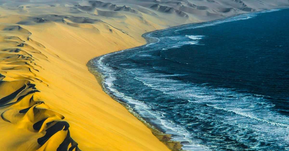 An aerial view of the orange dunes of Namib Sand Sea meeting the blue waves of the African coast.