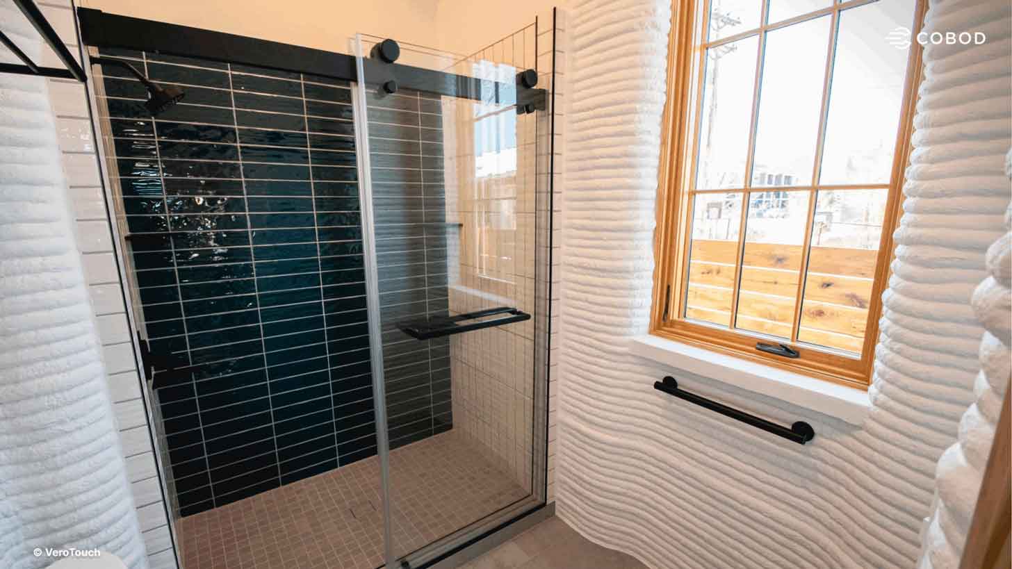 An interior view of a bathroom inside of a 3D-printed house, with layered walls and a clean tiled shower