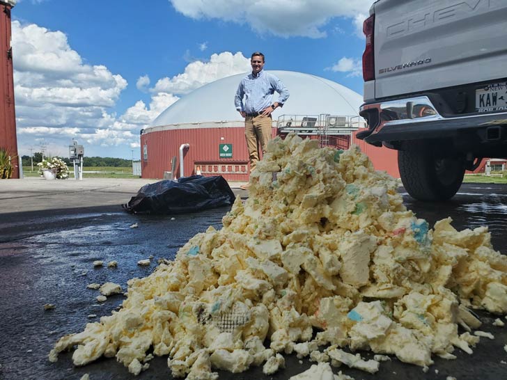 A pile of chunks of butter sits on the ground next to a truck. A man in a blue shirt and khakis stands looking at the pile, his hands on his hips