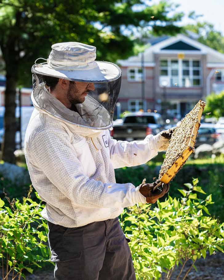 A man in a beekeeper's uniform holds up a hive in a lush green garden