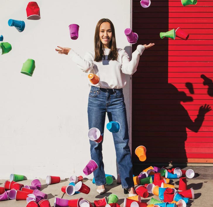 A college student, Lauren Choi, stands in front of a red and white wall, throwing colorful party cups in the air