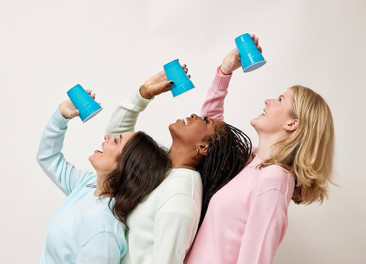 Three women stand in a row against a white backdrop. They wear pastel blue, green, and pink sweaters, and they hold up blue Hefty party cups over their mouths