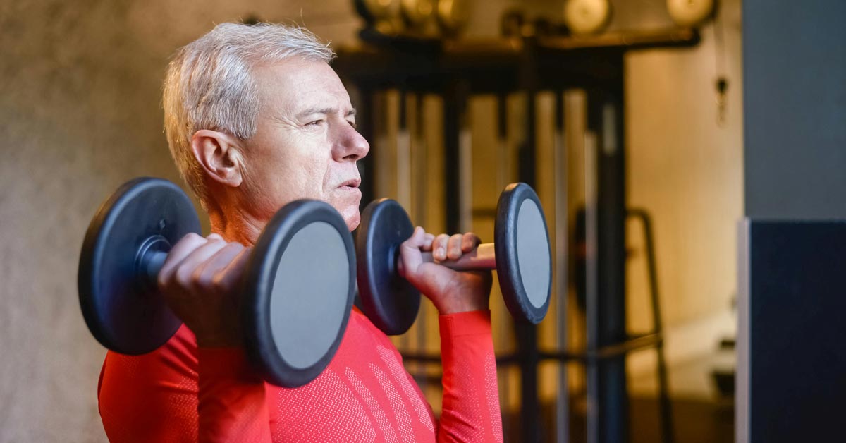 An older male adult in a red shirt lifts two dumbells in a gym