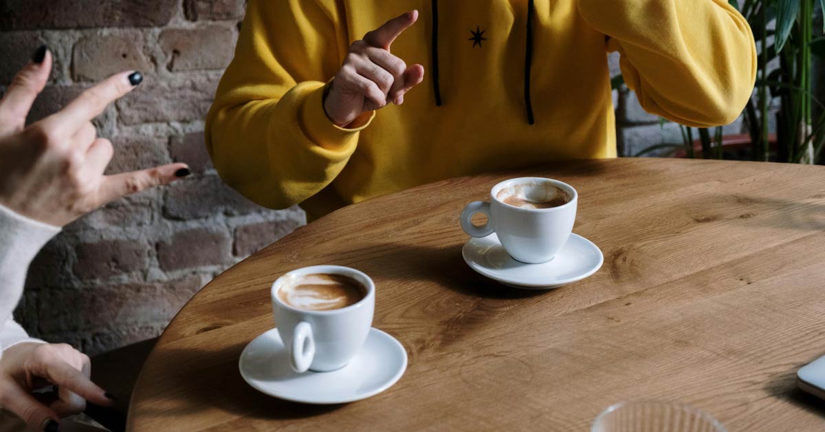 A close-up of two people communicating in sign language over two cups of coffee