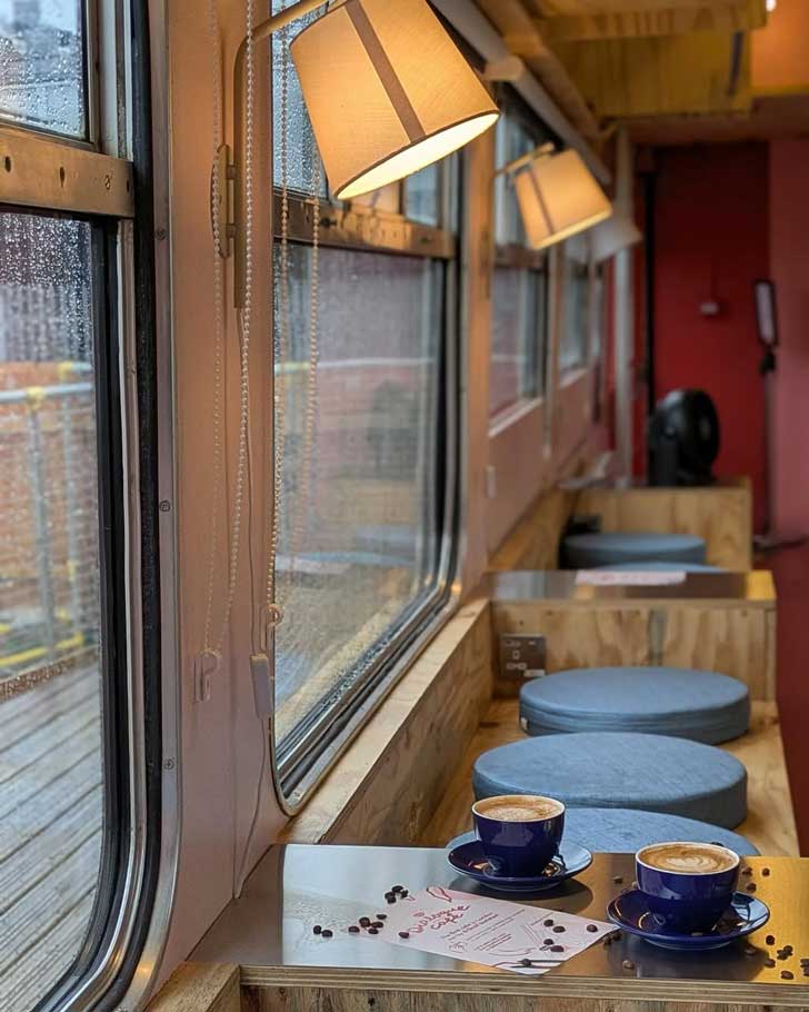 An interior view of a train car turned cafe, with slim wooden tables, warm lamps, and red walls
