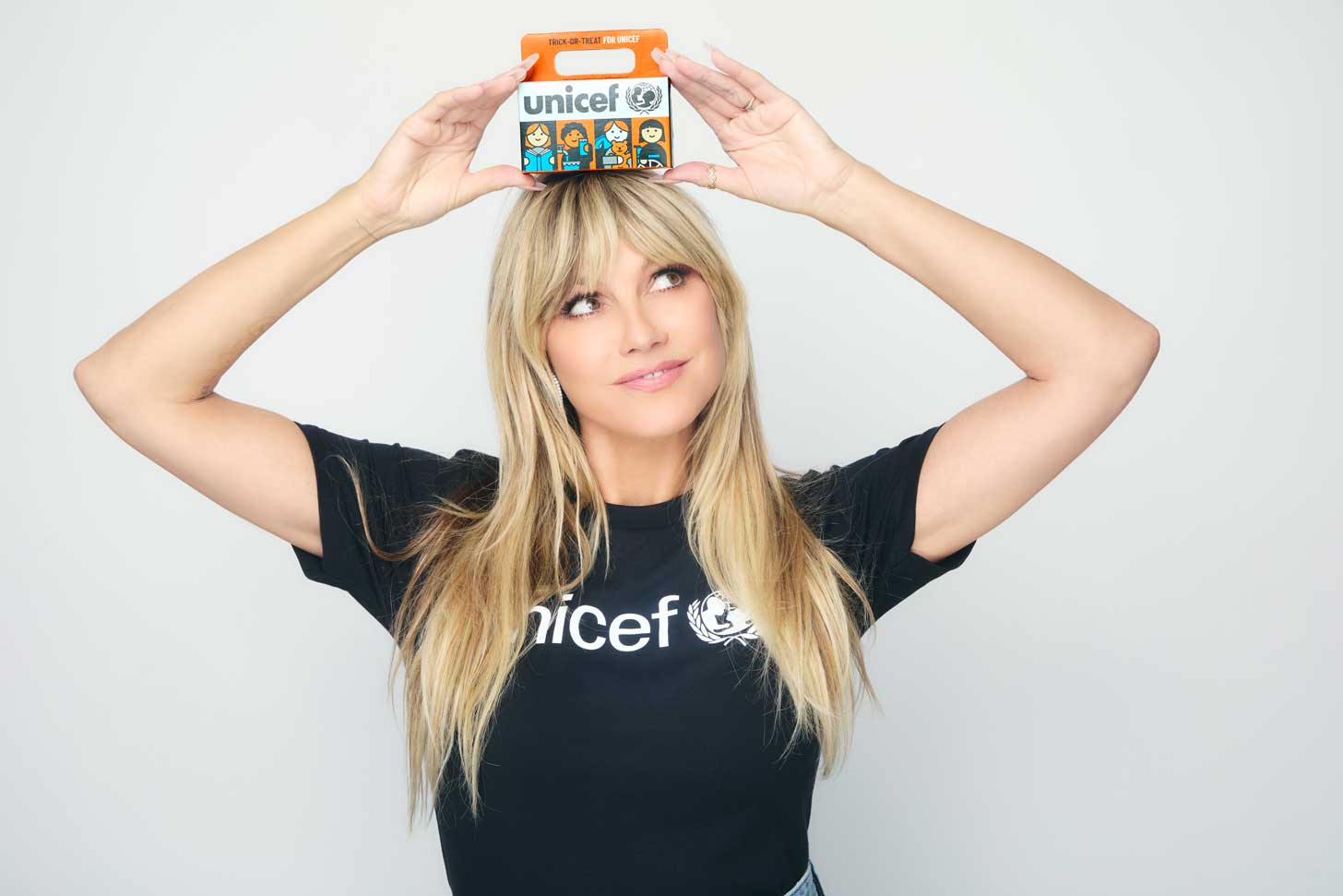 Heidi Klum wears a black t-shirt with the UNICEF logo on it, while balancing an orange box atop her head