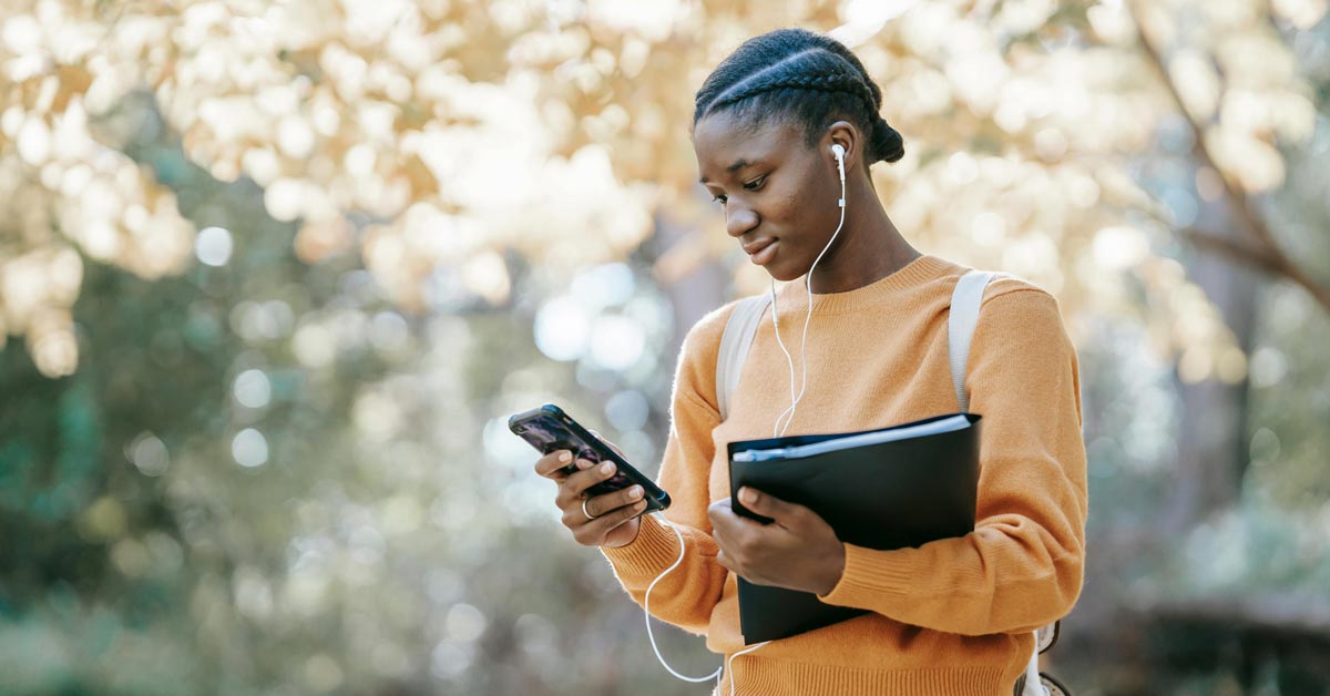 A Black teen girl in a yellow sweater stands outside looking at her phone. She has white wired earbuds in her ears.