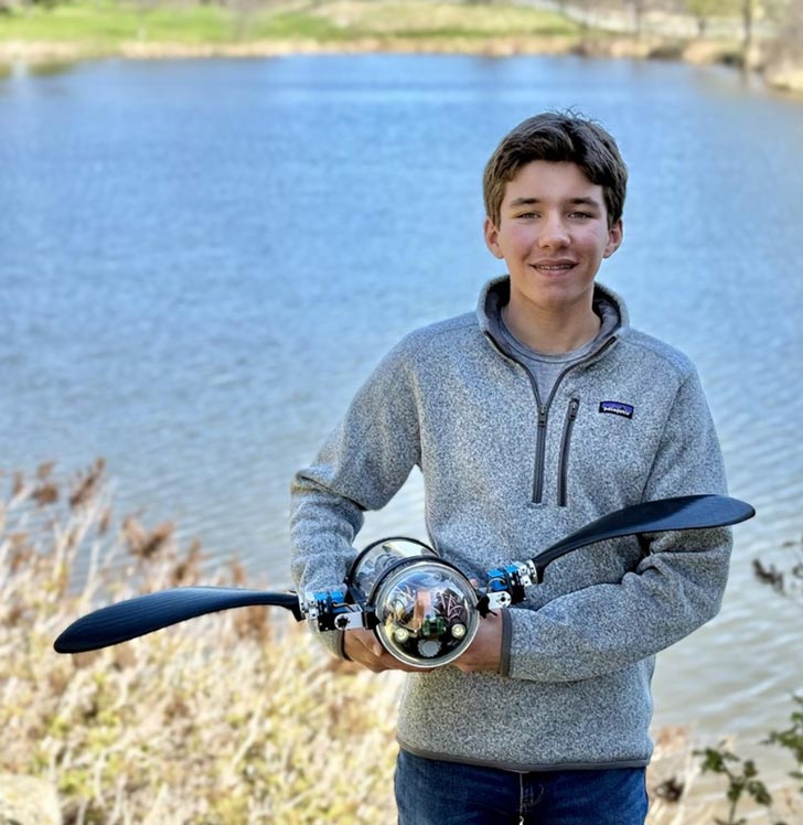 a 15-year-old boy holding up a robot and smiling in front of a lake